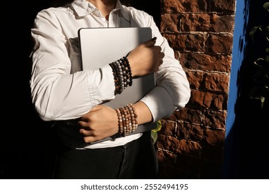 An unrecognizable girl in a white shirt holds a laptop computer in her hands against the background of a brick wall. There are bracelets made of stones on the hands. Close up. Place for text. 