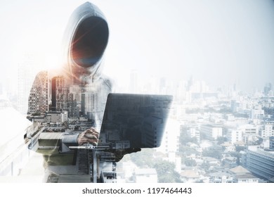 Unrecognizable hacker with hood using laptop on abstract city background. Hacking and malware concept. Double exposure 