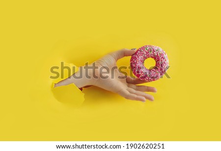 Unrecognizable hand holds delicious round glazed donut with sprinkle in torn yellow bright paper hole, popular food blog content, selective focus