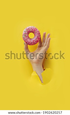 Unrecognizable hand holds delicious round glazed donut with sprinkle in torn yellow bright paper hole, vertical popular food blog content, selective focus