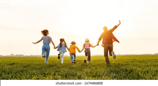 Unrecognizable happy   parents with daughters in casual clothes running back with holding hands while enjoying time together on green meadow in summer evening