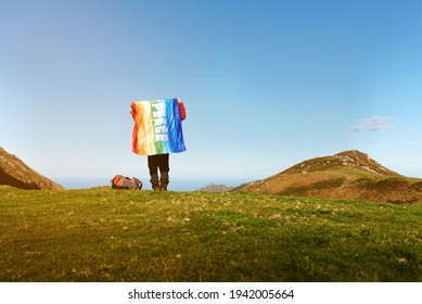 Unrecognizable Hiker With Backpack Holding A Rainbow Flag Of Peace On Top Of A Mountain On A Sunny Day. Person Making Summit. Day Of Peace.