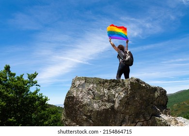 An Unrecognizable Hiker With A Backpack, Perched On A Rock At The Top Of A Mountain, Arms Raised Holding A Rainbow Flag Fluttering In The Wind. With A Blue Sky In The Background