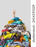 Unrecognizable human raises arm reaches out heap of colorful unfolded clothes busy doing wardrobe cleaning isolated over white background. Woman buried under cluttered clothing items. Decluttering