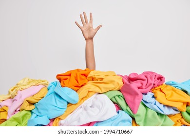 Unrecognizable human raises arm reaches out heap of colorful unfolded clothes busy doing wardrobe cleaning isolated over white background. Woman buried under cluttered clothing items. Decluttering - Shutterstock ID 1994710667
