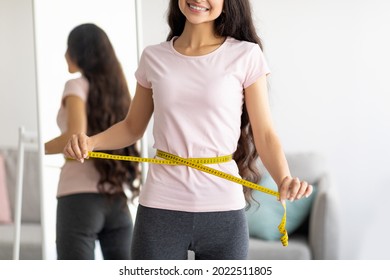 Unrecognizable Indian lady measuring her waist with tape in front of mirror at home, closeup. Cropped view of Asian woman checking her body parameters, losing weight indoors