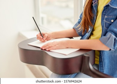 Unrecognizable Kid Writing Notes In Notepad On Acoustic Guitar While Sitting Near Window During Private Music Lesson In School