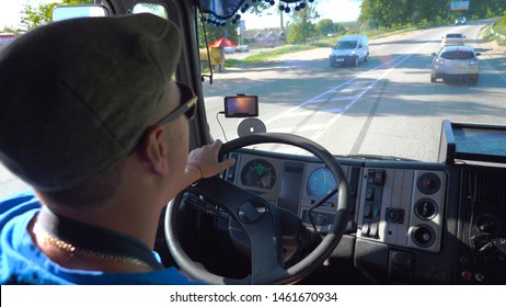 Unrecognizable Lorry Driver Riding On City Road At Sunny Day. Man In Cap And Sunglasses Controlling His Truck Attentive Watching Road. View Of Traffic From Inside Cabin. Inside Shot Slow Mo Close Up