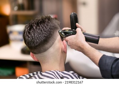 Unrecognizable male barber using electric razor to shave dark hair of man during work in barbershop
