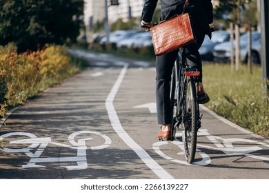 Unrecognizable male businessman in black suit riding on bicycle track in sleeping area of city. Crop view of office clerk with brown leather laptop bag using bike to get to job. Concept of activity. - Powered by Shutterstock