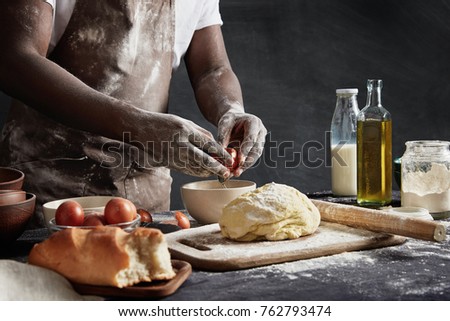 Unrecognizable male cook in apron, has dirty hands as prepares dough and mincemeat, cooks meat dumplings, breaks egg in bowl, isolated over black chalk background. Many ingridients on working table