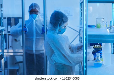 Unrecognizable Male Medical Scientist In Protective Workwear Opening Door To Laboratory Room, His Female Colleague Taking Test-tubes Off Shelf