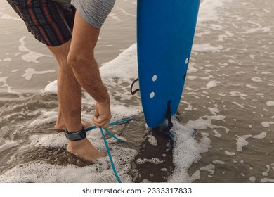 Unrecognizable male sportsman attaching leash of blue surfboard to ankle standing on sandy beach near sea. Man in striped shorts holding leg rope, preparing for activity on wet shore. - Powered by Shutterstock