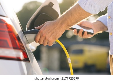 Unrecognizable male unplugging electric car from charging station. Man is unplugging in power cord to an electric car at sunset. Man charging electric car at charging station using smart phone app. - Powered by Shutterstock