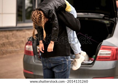 unrecognizable man in blacc clothes taking little teenage girl in car trunk, kidnapping children, rear view on maniac pedophile. on city street. violence, crime, children abuse concept. copy space