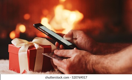 Unrecognizable Man Hands holding Credit Card and Using SmartPhone by the Burning Fireplace and Festive Presents - Close Up. Man with phone key-green screen by cozy fireside.