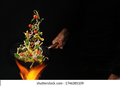 Unrecognizable Man Holding Wok Pan Above Fire And Cooking Pasta With Cherry Tomatoes, Onion And Basil Against Black Background. Close Up