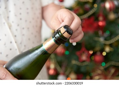 Unrecognizable Man Opening A Bottle Of Champagne Or Cider With A Christmas Tree In The Background. Concept Of Celebratory Toast. Close Up Image With Copy Space.