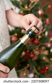 Unrecognizable Man Opening A Bottle Of Champagne Or Cider With A Christmas Tree In The Background. Concept Of Celebratory Toast. Close Up Image With Copy Space.