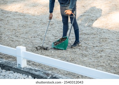 unrecognizable man is seen using a rake and scoop to clean up horse manure in an equestrian center. scene reflects the often unseen but essential work involved in maintaining clean and healthy stable - Powered by Shutterstock