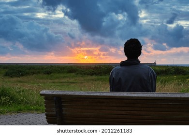 Unrecognizable Man Sitting On A Bench Enjoying In Solitude A Beautiful Sunset With A Dramatic Sky Full Of Clouds And Color.