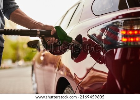 Unrecognizable man using hose with nozzle to fill fuel tank of red car with gasoline in daytime at station