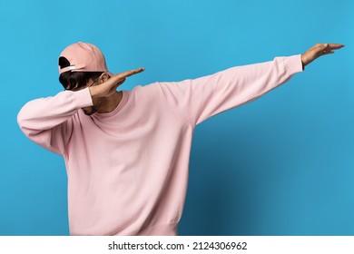Unrecognizable millennial guy in pink sweatshirt and cap danicng hip hop alone on blue studio background, dabbing with his hands, covering face with arm, panorama with copy space