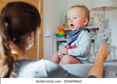 Unrecognizable mother pulling hands to her one years old infant boy. Little baby sitting on the bed and communication with his mother in cozy home interior.
