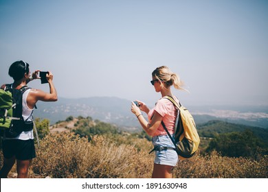 Unrecognizable multiethnic travelers wearing summer clothes and sunglasses with backpacks standing on hill and photographing landscape with mobile phone in sunny day