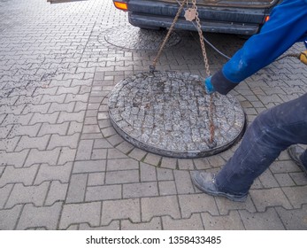 Unrecognizable Municipal Utility Worker Opening A Heavy Double Manhole Cover On A Road Paved With Concrete Paving Stones.