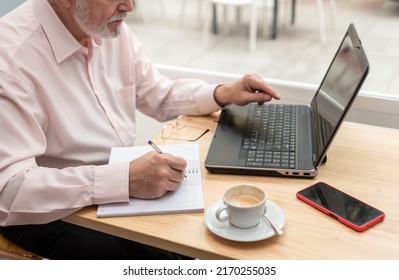 An Unrecognizable Older Man Sitting In A Coffee Shop Or Bar Studying And Using His Laptop To Make Notes