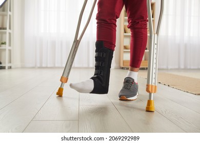 Unrecognizable person with broken leg or foot injury walking on crutches. Man wearing leg brace ankle support adjustable strap fracture fixator standing in living room. Cropped low section close up