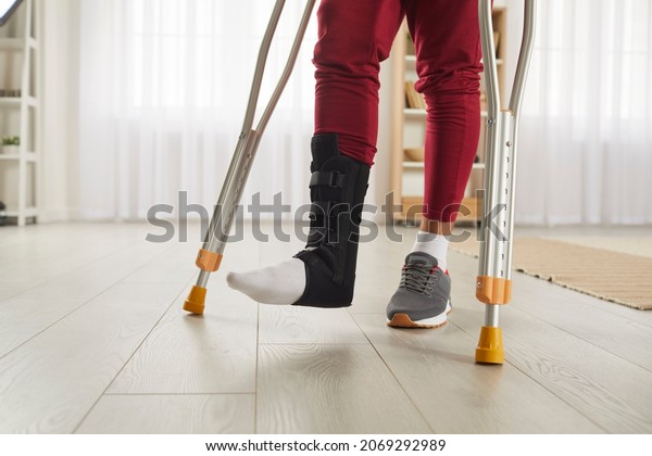 Unrecognizable person with broken leg or foot injury\
walking on crutches. Man wearing leg brace ankle support adjustable\
strap fracture fixator standing in living room. Cropped low section\
close up