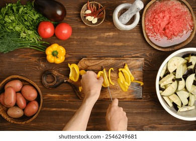 Unrecognizable person chopping bell peppers on a wooden cutting board. Bright yellow peppers, tomatoes, and eggplants are ready for a delicious home-cooked meal. - Powered by Shutterstock