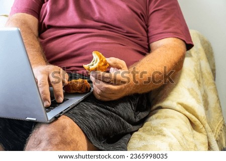 Unrecognizable person. Sedentary overweight man, sitting eating pastries next to a laptop.