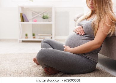 Unrecognizable pregnant woman caressing her belly at home. Young expectant blonde feeling her baby push, sitting on floor, copy space, side view. Pregnancy, rest, life, expectation concept