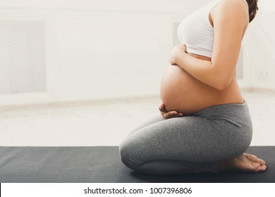 Unrecognizable pregnant woman training yoga in hero pose, caressing her belly. Young happy expectant relaxing, thinking about her baby and enjoying her future life. Motherhood, pregnancy, yoga concept