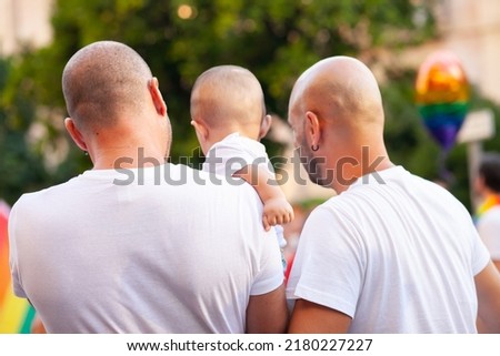 Unrecognizable real people from behind at Pride parade, support for LGBTQIA community. Two gay fathers with child from behind. Gay family. Crowd of people at public event. Lgbtq+  parents.