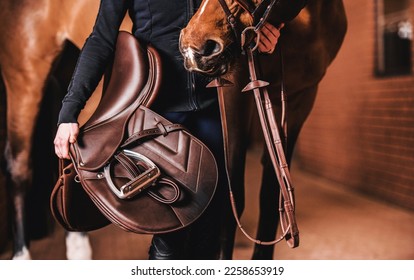 Unrecognizable Rider Standing Next to the Horse Inside Modern Red Brick Stable with Brown Leather Saddle in the Hand. Equestrian Lifestyle Theme. - Powered by Shutterstock
