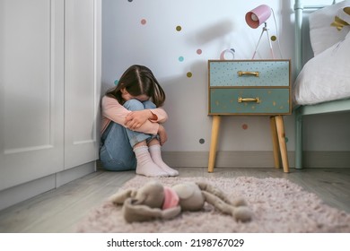 Unrecognizable Sad Little Girl Sitting On The Floor Of Her Bedroom With Stuffed Toy Lying. Selective Focus On Girl In Background