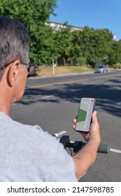 Unrecognizable Senior Man Looking At An Online Map In An App On His Phone Before Riding His Electric Kick Scooter In A Sunny City. Concepts: Use Of Technology By The Elderly, Active Retired Life.