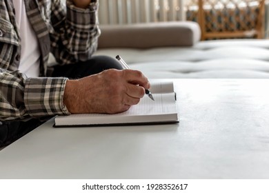 Unrecognizable Senior Man Writing In His Book In The Lounge At Home. Close Up Of An Older Man Writing Something Down On A Diary With Pen. Old Person's Hands Holding A Pen And Making Notes In A Diary.