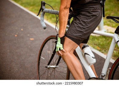 Unrecognizable Senior Woman Cyclist Holding Hand On Knee In Pain, Massaging Her Gliding Joint Bone. Arthritis Cramp In The Knee While Cycling Outdoor. Copy Space