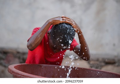 Unrecognizable Small Black African Village Boy Kneeling In Front Of A Brown Plastic Tub Vigorously Washing His Hair Splashing Water Aroung