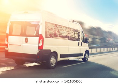 Unrecognizable small passenger van hurry up on highway at city street traffic with urban cityscape and sunset sky on background. Charter or shuttle bus  van hurry up on road with motion blur effect - Shutterstock ID 1411804553