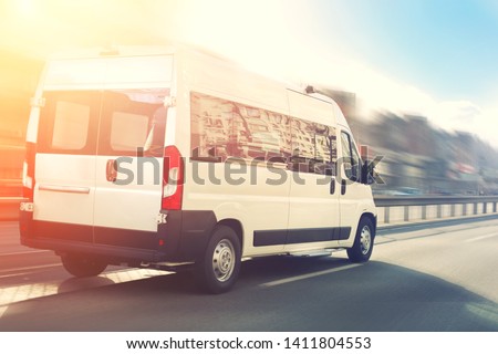 Unrecognizable small passenger van hurry up on highway at city street traffic with urban cityscape and sunset sky on background. Charter or shuttle bus  van hurry up on road with motion blur effect