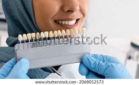 Unrecognizable Stomatologist In Blue Gloves Holding Dental Teeth Shade Guide Chart, Choosing Right Enamel Color For Muslim Female Patient In Hijab During Stomatologic Treatment In Clinic, Closeup