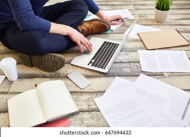 Unrecognizable student  sitting cross legged on wooden floor, using laptop computer while studying and preparing for exams