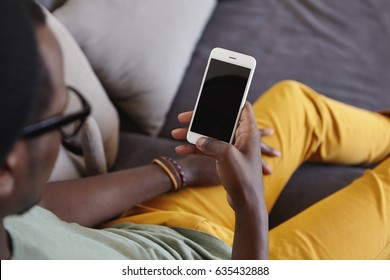 Unrecognizable stylish Afro American man wearing yellow jeans relaxing at home, lying on comfortable sofa, reading message on smart phone with black blank copy space screen for your information