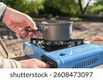 Unrecognizable traveler cooking outdoors while camping at a nature campground a sunny morning. Close up view, one hand lights the blue portable stove, the other one holds a pot on it.